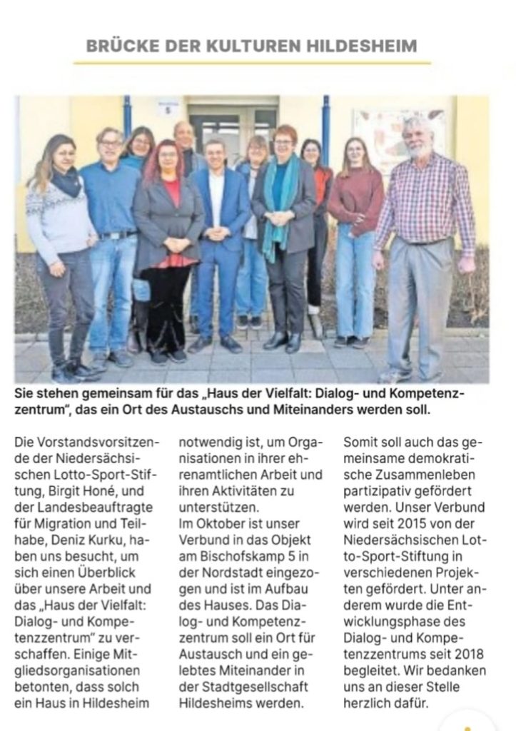 Das Bild zeigt eine Gruppe von elf Erwachsenen, die in zwei Reihen stehen und in die Kamera lächeln. Im Hintergrund ist ein hellgraues Gebäude mit Fenstern und Türen zu sehen. Der Text neben dem Bild beschreibt, dass diese Personen gemeinsam für das "Haus der Vielfalt: Dialog- und Kompetenzzentrum" stehen, das ein Ort des Austauschs und Miteinanders werden soll. Es wird erwähnt, dass das Haus der Vielfalt seit 2015 von der Niedersächsischen Lotto-Sport-Stiftung gefördert wird.