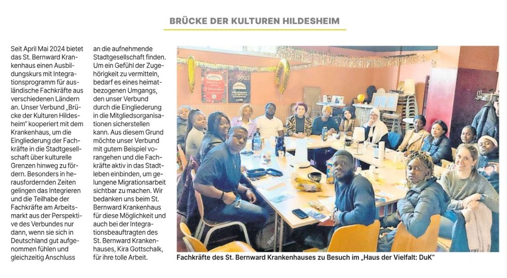 Das Bild zeigt eine Gruppe von etwa 15 bis 20 Personen, die an einem runden Tisch in einem hellen Raum sitzen und sich unterhalten. Im Hintergrund sind Regale mit Büchern und verschiedenen Objekten zu erkennen, darunter ein gelbes Plakat mit dem Jahreszahlen "2023" und "2024". Der Text neben dem Bild erklärt, dass das St. Bernward Krankenhaus seit April 2024 einen Ausbildungskurs mit Integrationsprogramm für ausländische Fachkräfte anbietet und dafür mit dem Verbund "Brücke der Kulturen Hildesheim" kooperiert. Das Foto wurde bei einem Besuch der Fachkräfte des St. Bernward Krankenhauses im "Haus der Vielfalt: DuK" aufgenommen.