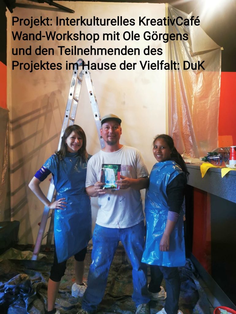 Ein Foto von einem "Wand-Workshop" des Projekts "Interkulturelles KreativCafé" im Haus der Vielfalt: DuK. In der Bildmitte steht Ole Görgens, ein Mann mittleren Alters mit einem Bart, der ein T-Shirt mit einem Bild eines Wasserfalls trägt und ein kleines, gerahmtes Kunstwerk in den Händen hält. Zu seiner Linken steht eine lächelnde junge Frau mit langen, dunklen Haaren, die eine blaue Schürze trägt. Zu seiner Rechten steht eine weitere junge Frau mit dunklen Haaren und einem Lächeln, ebenfalls in einer blauen Schürze. Alle drei Personen tragen Arbeitsschuhe. Im Hintergrund sind Malerleitern und mit Folie abgedeckte Wände zu sehen, was auf eine laufende Renovierungs- oder Gestaltungstätigkeit hindeutet.