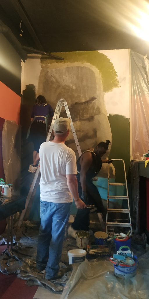 Das Foto zeigt drei Personen, die gemeinsam an einer großen Wand arbeiten, die teilweise bereits mit einer neuen Farbschicht versehen ist. Die Wand weist eine organische Form auf, die an eine Felsformation oder einen Wasserfall erinnert und in verschiedenen Grau- und Grüntönen gehalten ist. Eine Person in einem weißen T-Shirt und Jeans steht im Vordergrund mit dem Rücken zum Betrachter und schaut auf die Wand. Links im Bild steht eine Person auf einer Leiter und arbeitet am oberen Teil der Wand. Rechts steht eine weitere Person in dunkler Kleidung ebenfalls auf einer Leiter und konzentriert sich auf den unteren Bereich der Wand. Überall auf dem Boden liegen Schutzplanen, und es sind verschiedene Malutensilien wie Farbeimer und Pinsel zu sehen, was auf eine aktive Gestaltungssession hinweist.
