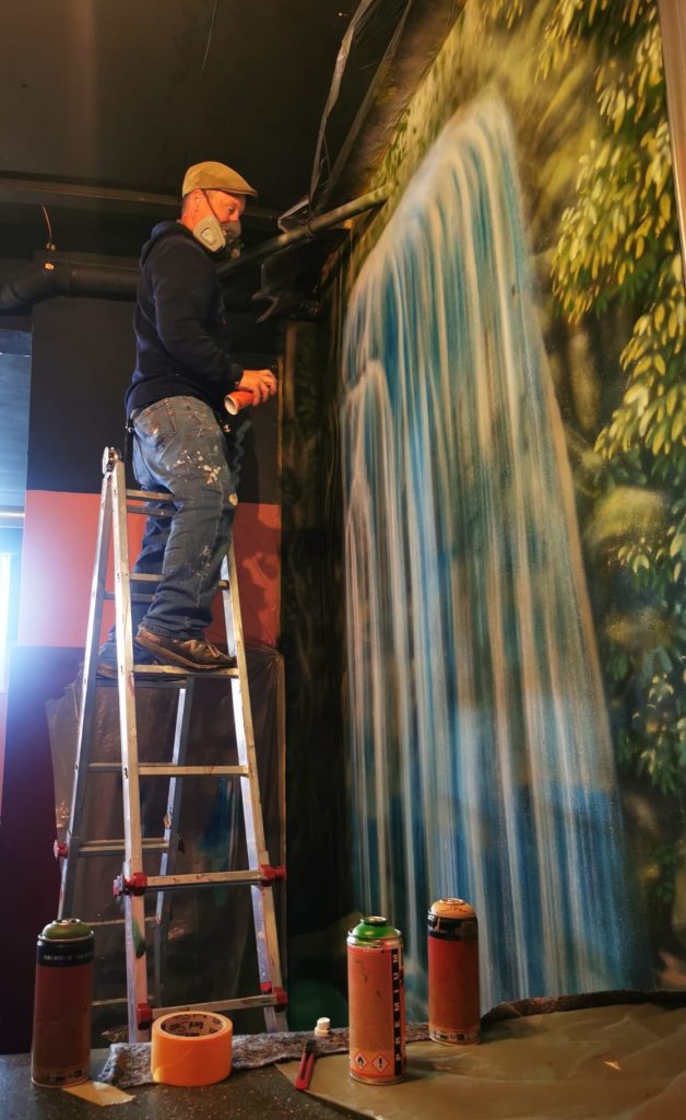 Dieses Bild zeigt einen Mann, der auf einer Leiter steht und mit einer Sprühdose ein Wandgemälde gestaltet, das einen Wasserfall und üppiges Grün darstellt. Er trägt eine Schutzausrüstung, einschließlich einer Gesichtsmaske, und ist in einem grauen T-Shirt, Jeans und einer Mütze gekleidet. Unter der Leiter und auf dem Boden sind Farbdosen, Malerband und weitere Materialien zu sehen. Das Wandgemälde nimmt fast die gesamte Höhe der Wand ein und ist sehr detailliert.
