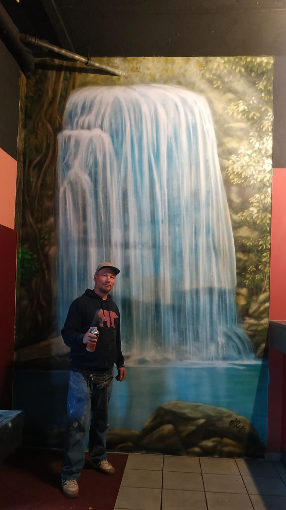 Das Bild zeigt ein großes, detailliertes Wandgemälde eines Wasserfalls, der eine Wand hinunterstürzt, umgeben von üppiger grüner Vegetation. Ein großer, realistisch gemalter Baumstamm mit Ästen ist auf der rechten Seite des Bildes zu sehen. Die Farben sind lebendig und detailliert, besonders die blauen und weißen Farbtöne des Wassers, die das fließende Element hervorheben